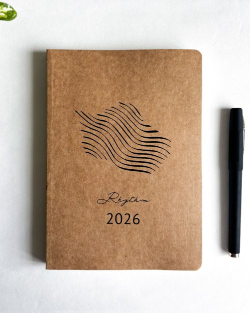 The Rhythm 2026 planner featuring its natural brown Kraft paper cover and subtle wave motif, alongside a pen and earbuds, representing its high-quality, eco-friendly materials and minimalist aesthetic for mindful organization