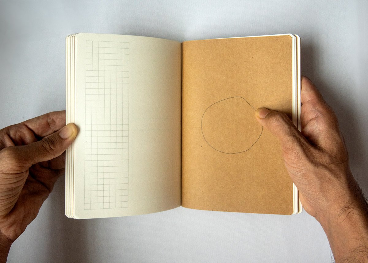 Hands holding an open journal featuring a vertical grid column on the left and a simple circular motif on a kraft paper right page, illustrating a flexible layout for gentle daily planning