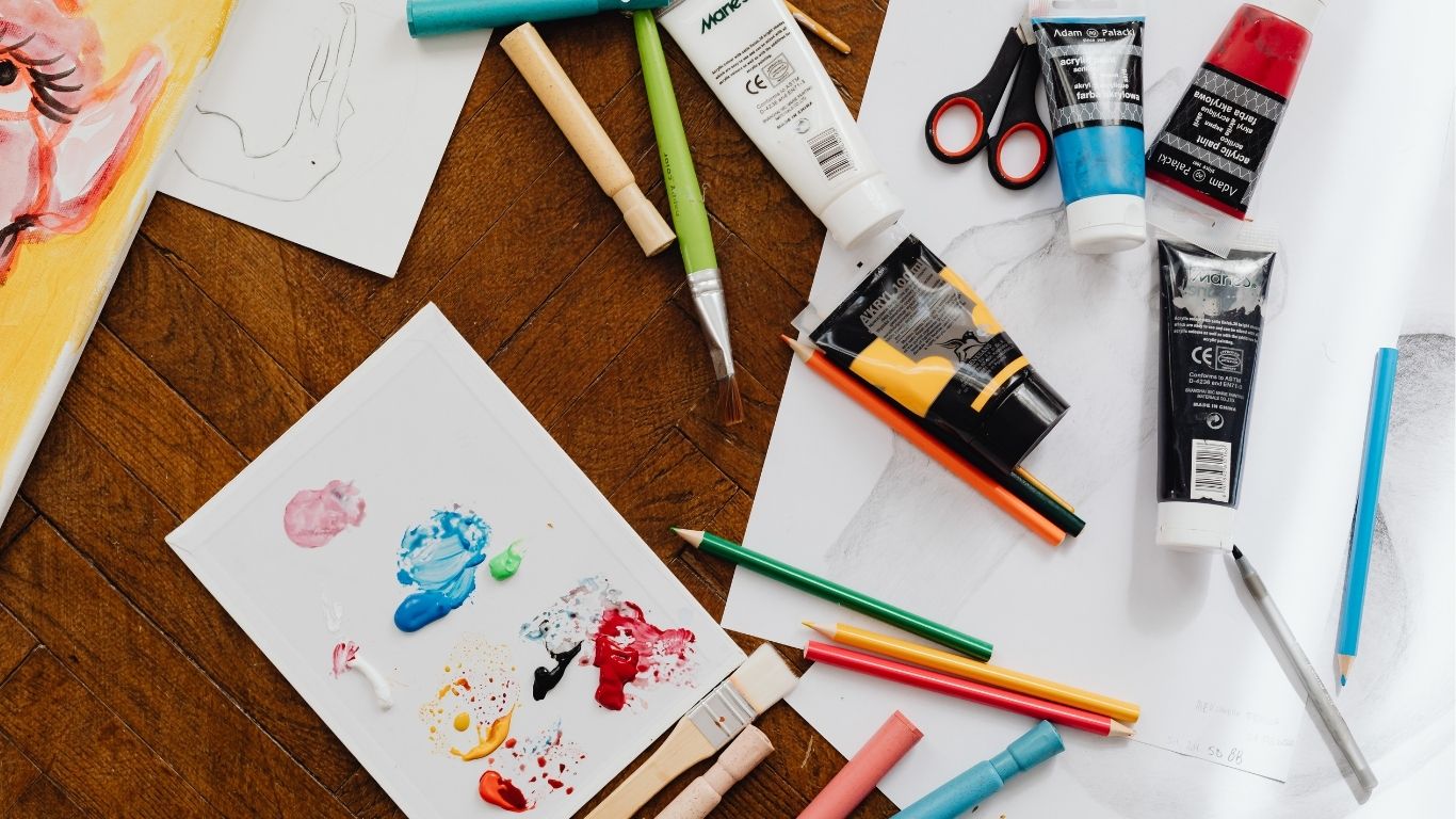 Art supplies scattered on a wooden table including watercolors, colored pencils, chalk pastels, and paper with colorful paint splotches