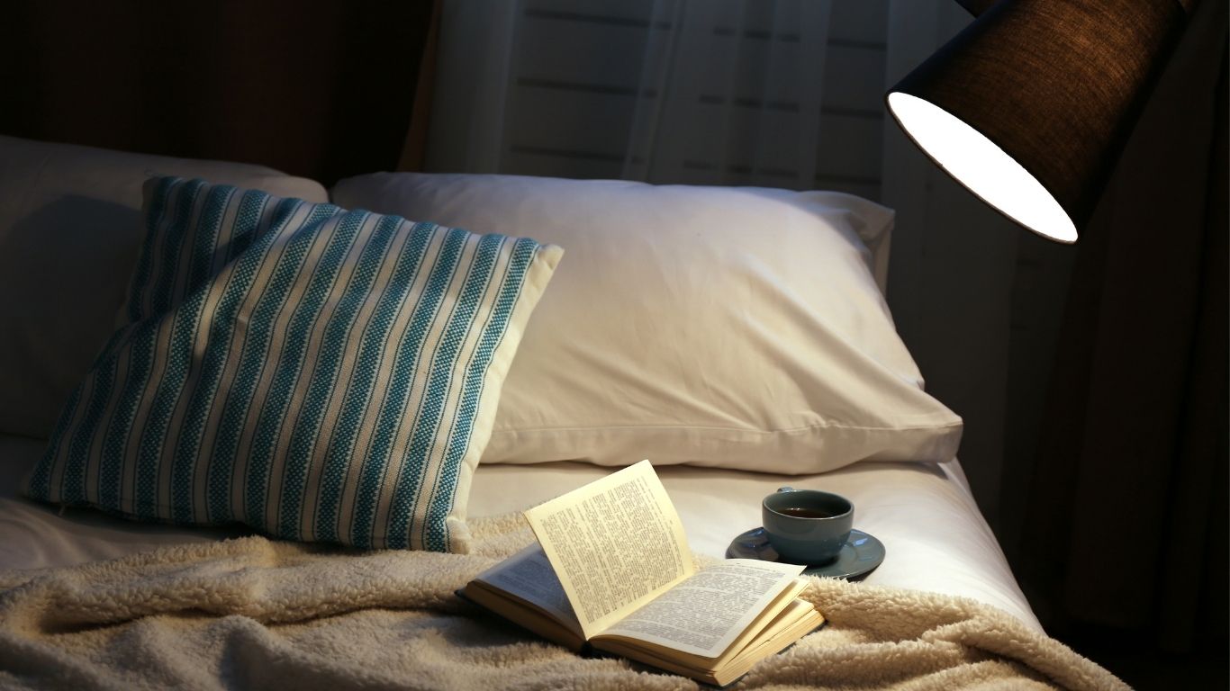 Cozy bed with an open book, a cup of tea, striped pillows, and a warm bedside lamp